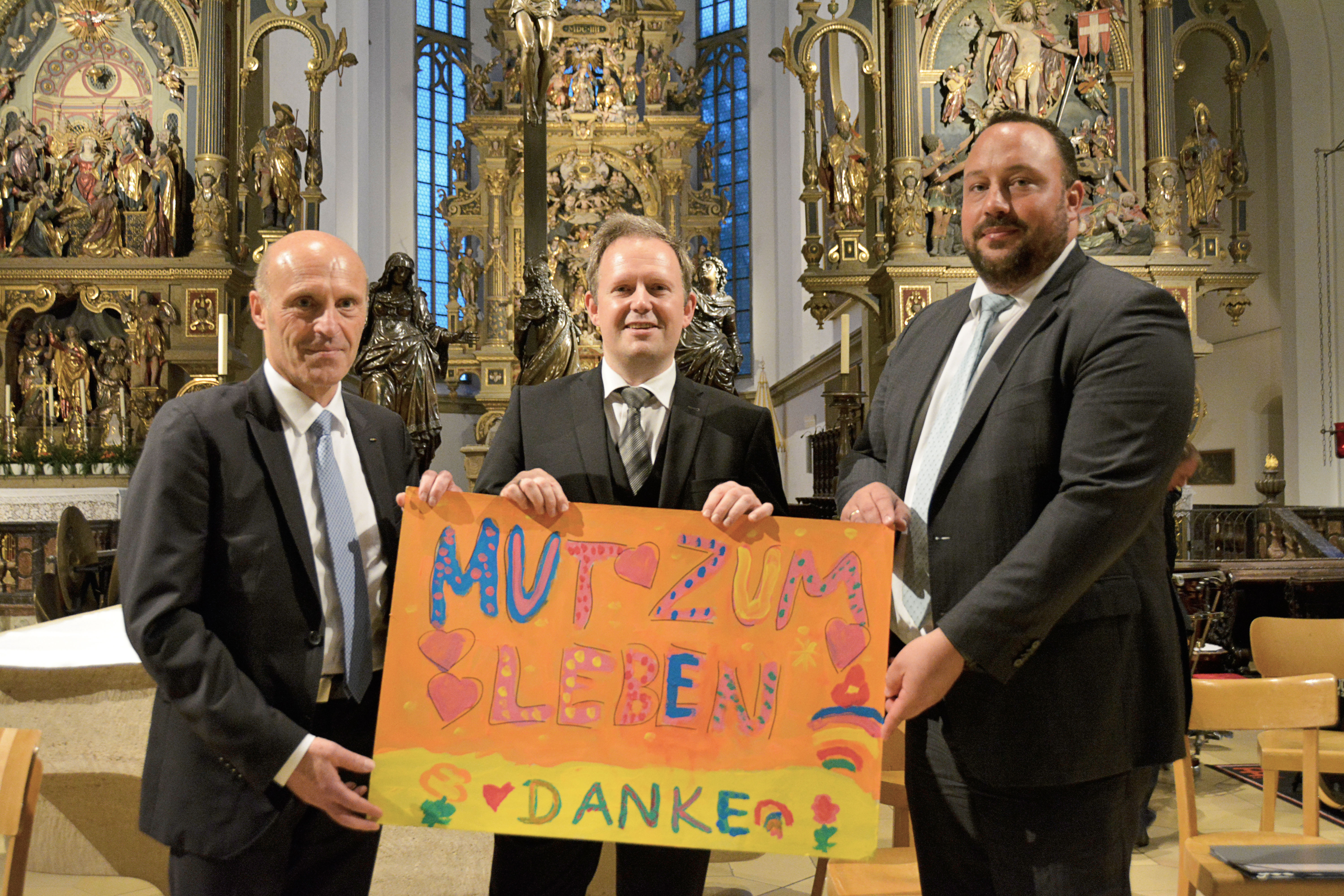 Markus Mayer, Vorstandsvorsitzender der KJF Augsburg (links), überreichte gemeinsam mit Daniel Kiesel Vorstand Soziales und stellvertretender Vorstandsvorsitzender der KJF Augsburg (rechts), dem Dirigenten Bernhard Willer ein von Kindern und Jugendlichen des Frère-Roger-Kinderzentrums gestaltetes Plakat. Foto: KJF Augsburg / Jasmin Mächtlinger